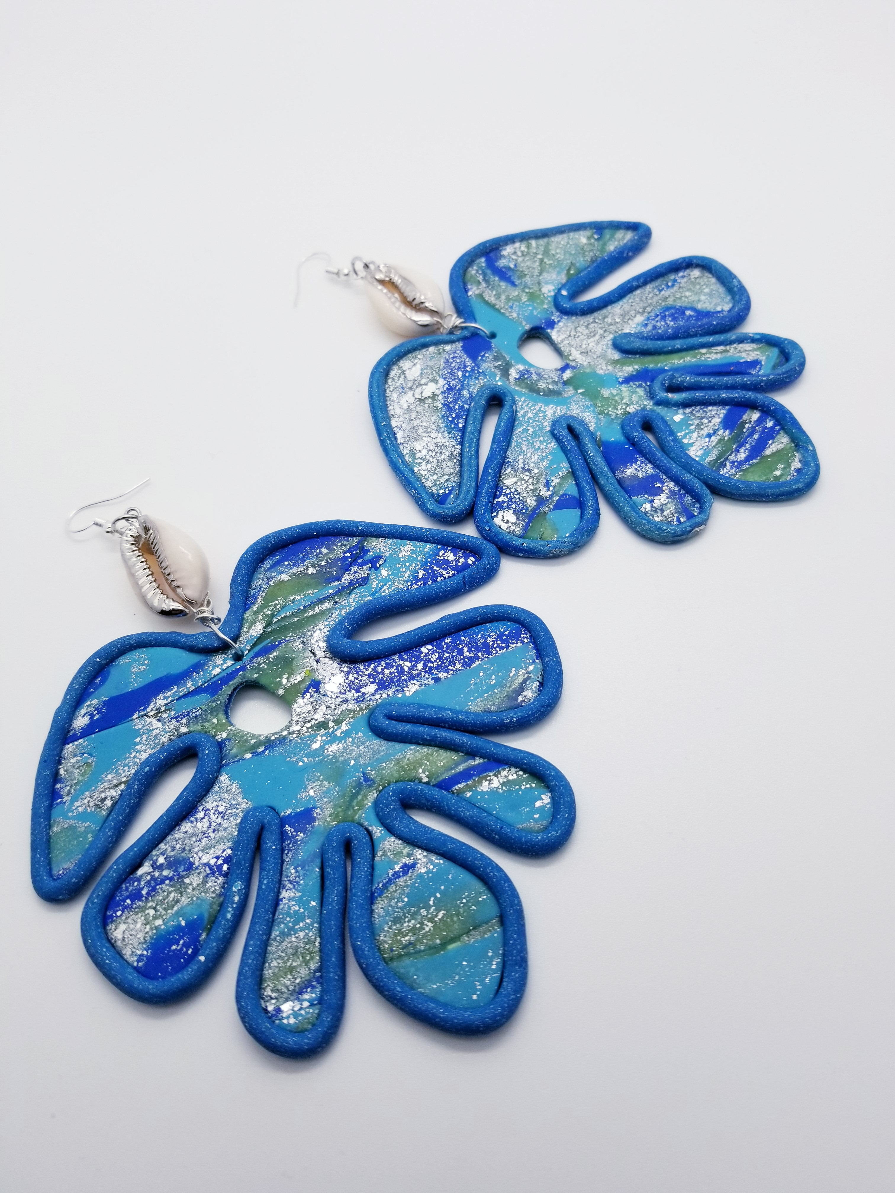 Length: 5 inches | Width: 3.5 inches | Weight: 1.5 ounces The Monstera plant represents Honor, Respect, and Longevity! These earrings are handmade using blue marble polymer clay, silver flakes, and ceramic cowrie shell on silver alloy hypoallergenic hook with rubber back closures.